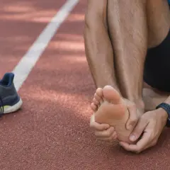 Atleta seduto su una pista, si tiene il piede destro, lamentando un forte bruciore ai piedi dopo la corsa. Scarpe da ginnastica a sinistra.