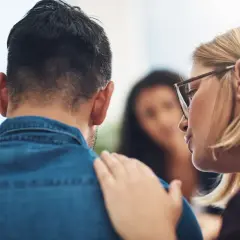 Una donna con gli occhiali consola un uomo, offrendo supporto per la psicosi da stress. Un'altra persona è sfocata sullo sfondo.