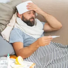 Uomo a letto con termometro e panno umido sulla fronte, cerca consigli su come abbassare la febbre. Sul comodino, limone e bicchiere d'acqua.