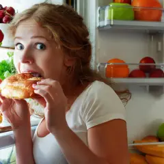 Donna con occhi spalancati mangia un panino aperto dal frigorifero pieno di frutta e verdura, sembra avere sempre fame.