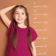 A young girl with braided hair smiles as she measures her height against a chalk growth chart.