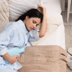 Donna a letto con una borsa dell'acqua calda sull'addome, cercando sollievo. Un'immagine che evoca il desiderio di sapere come far venire il ciclo.