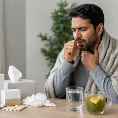 Uomo malato tossisce, coperto da una coperta, con sul tavolo farmaci, tra cui brufen e tachipirina insieme, tisana e fazzoletti.
