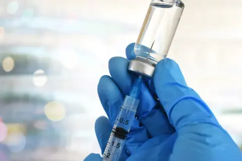 Mano guantata blu aspira liquido da fiala. Preparazione vaccino herpes zoster, attenzione agli effetti collaterali.