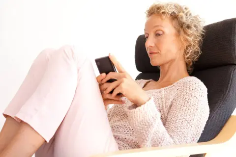 Donna bionda con capelli ricci, seduta su una poltrona, guarda il suo smartphone. Un'immagine che ispira su come superare una separazione a 50 anni.