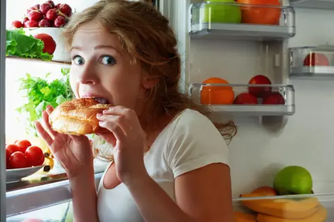 Donna con occhi spalancati mangia un panino aperto dal frigorifero pieno di frutta e verdura, sembra avere sempre fame.