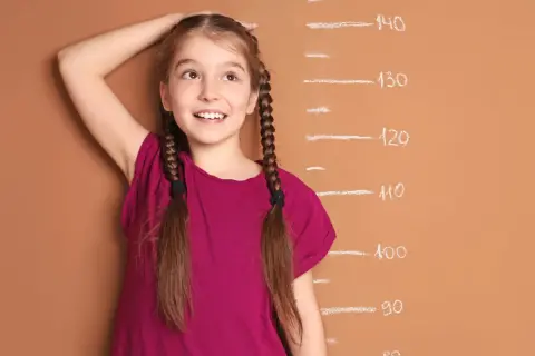 A young girl with braided hair smiles as she measures her height against a chalk growth chart.