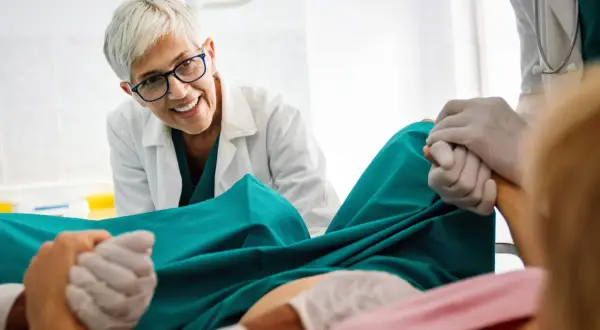 Medico sorridente assiste a un parto, tenendo la mano della paziente. Si percepisce un momento di scollamento membrane e attesa.
