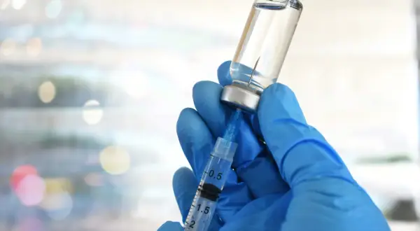 Mano guantata blu aspira liquido da fiala. Preparazione vaccino herpes zoster, attenzione agli effetti collaterali.