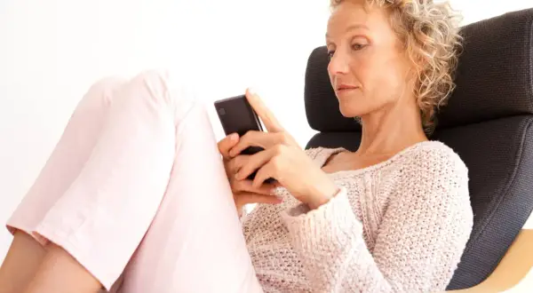 Donna bionda con capelli ricci, seduta su una poltrona, guarda il suo smartphone. Un'immagine che ispira su come superare una separazione a 50 anni.