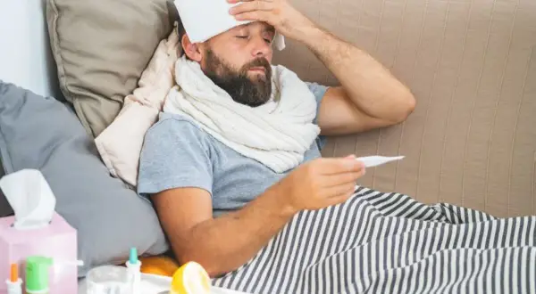 Uomo a letto con termometro e panno umido sulla fronte, cerca consigli su come abbassare la febbre. Sul comodino, limone e bicchiere d'acqua.