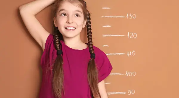 A young girl with braided hair smiles as she measures her height against a chalk growth chart.