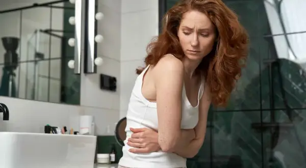 Donna con capelli rossi in bagno, stringe l'addome con le mani, visibilmente sofferente. Potrebbe essere un caso di diarrea prima del ciclo.