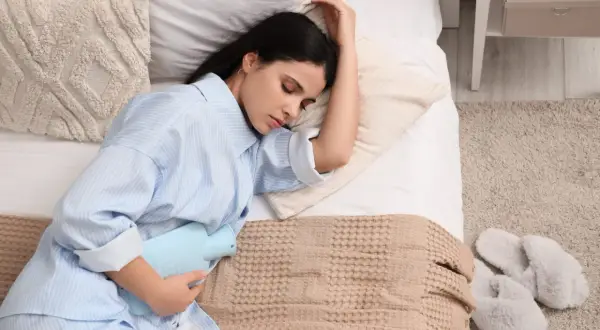 Donna a letto con una borsa dell'acqua calda sull'addome, cercando sollievo. Un'immagine che evoca il desiderio di sapere come far venire il ciclo.
