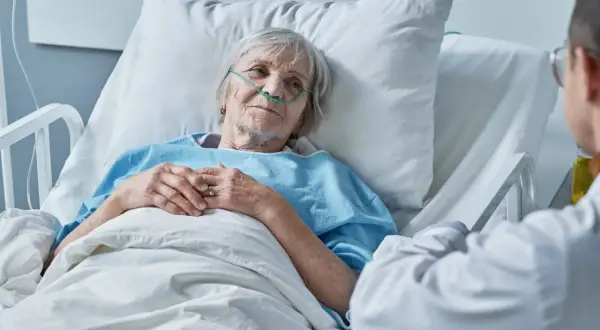Anziana paziente in ospedale con cannula nasale, che sembra accettare la morte con serenità, mentre un medico le parla.