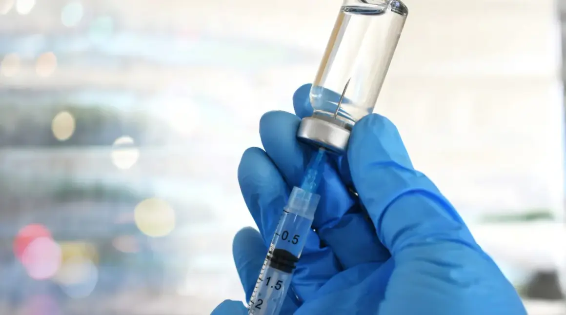 Mano guantata blu aspira liquido da fiala. Preparazione vaccino herpes zoster, attenzione agli effetti collaterali.