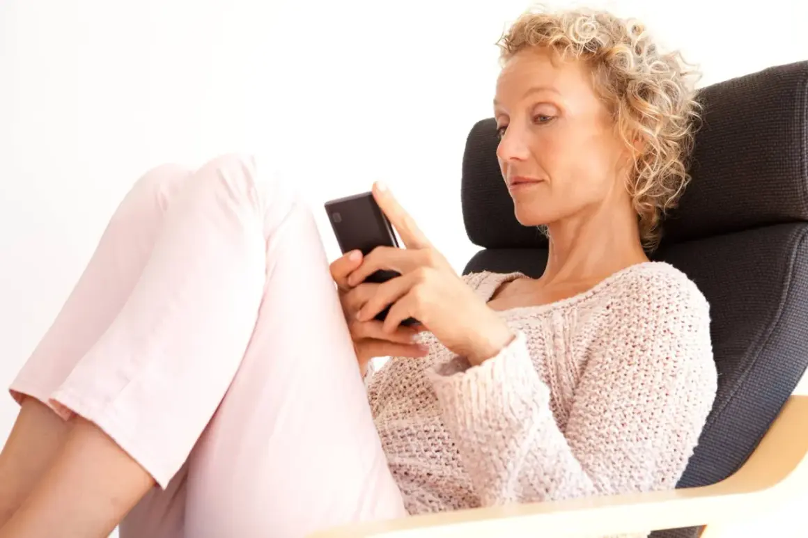 Donna bionda con capelli ricci, seduta su una poltrona, guarda il suo smartphone. Un'immagine che ispira su come superare una separazione a 50 anni.