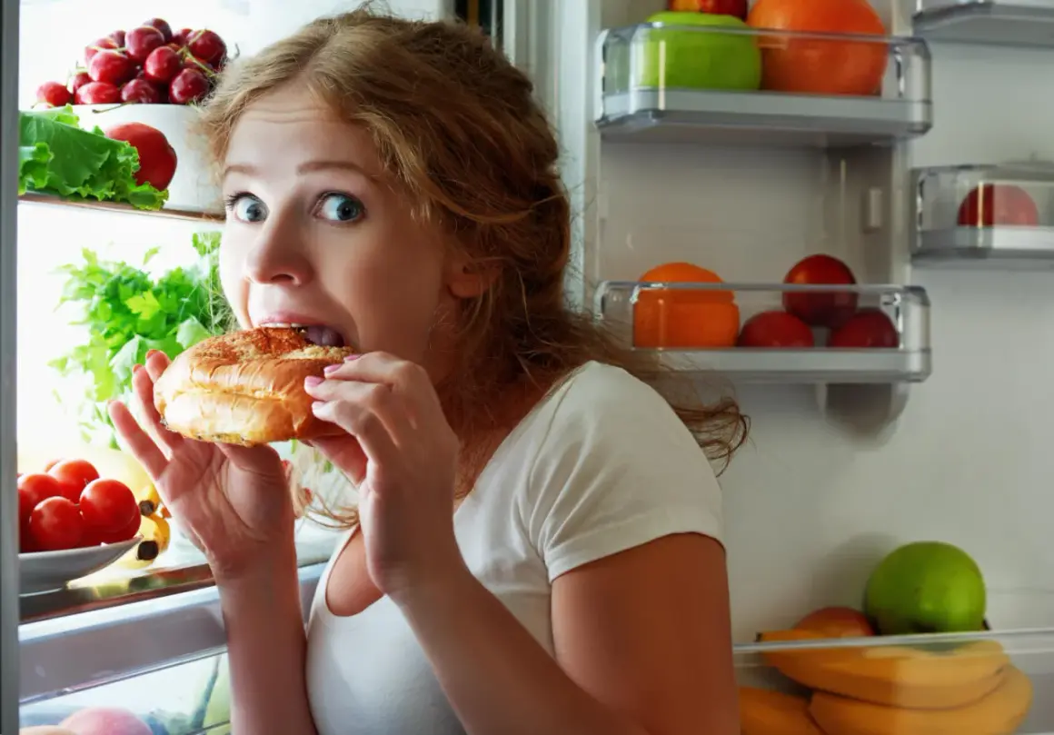 Donna con occhi spalancati mangia un panino aperto dal frigorifero pieno di frutta e verdura, sembra avere sempre fame.