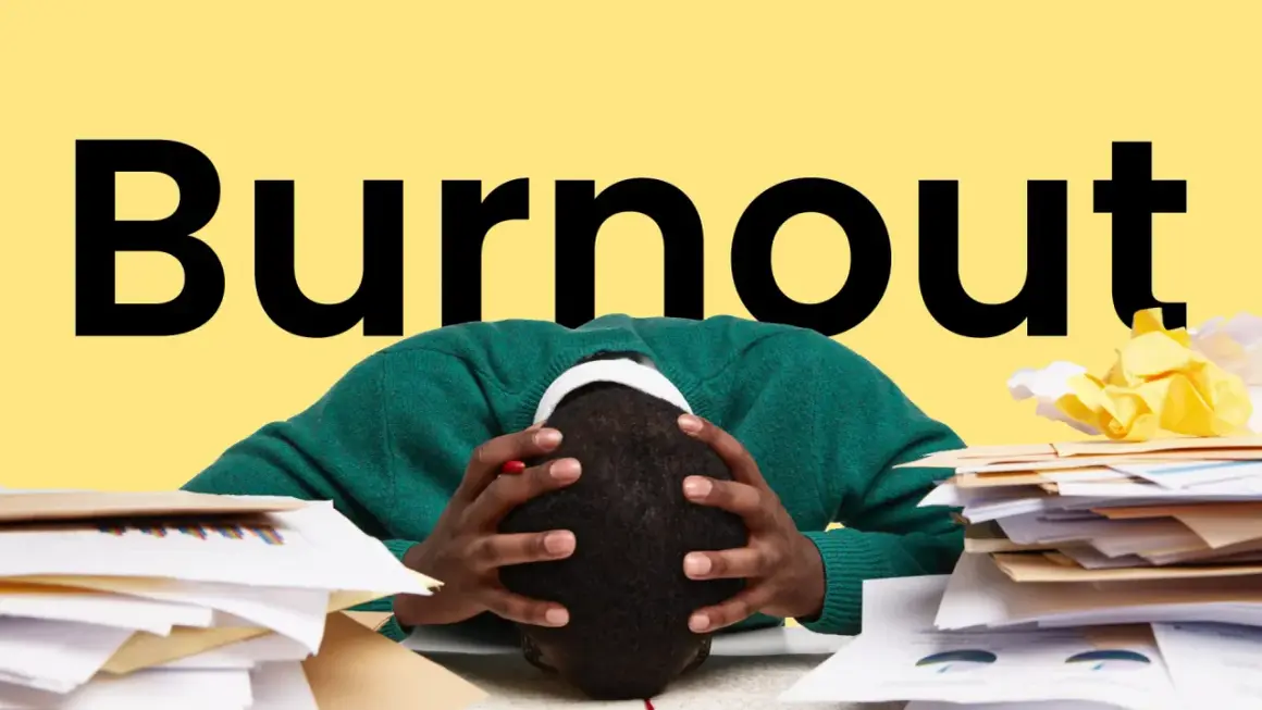 Persona con la testa tra le mani, circondata da pile di documenti, con la parola "Burnout" in grande sullo sfondo giallo. Sintomi di esaurimento.