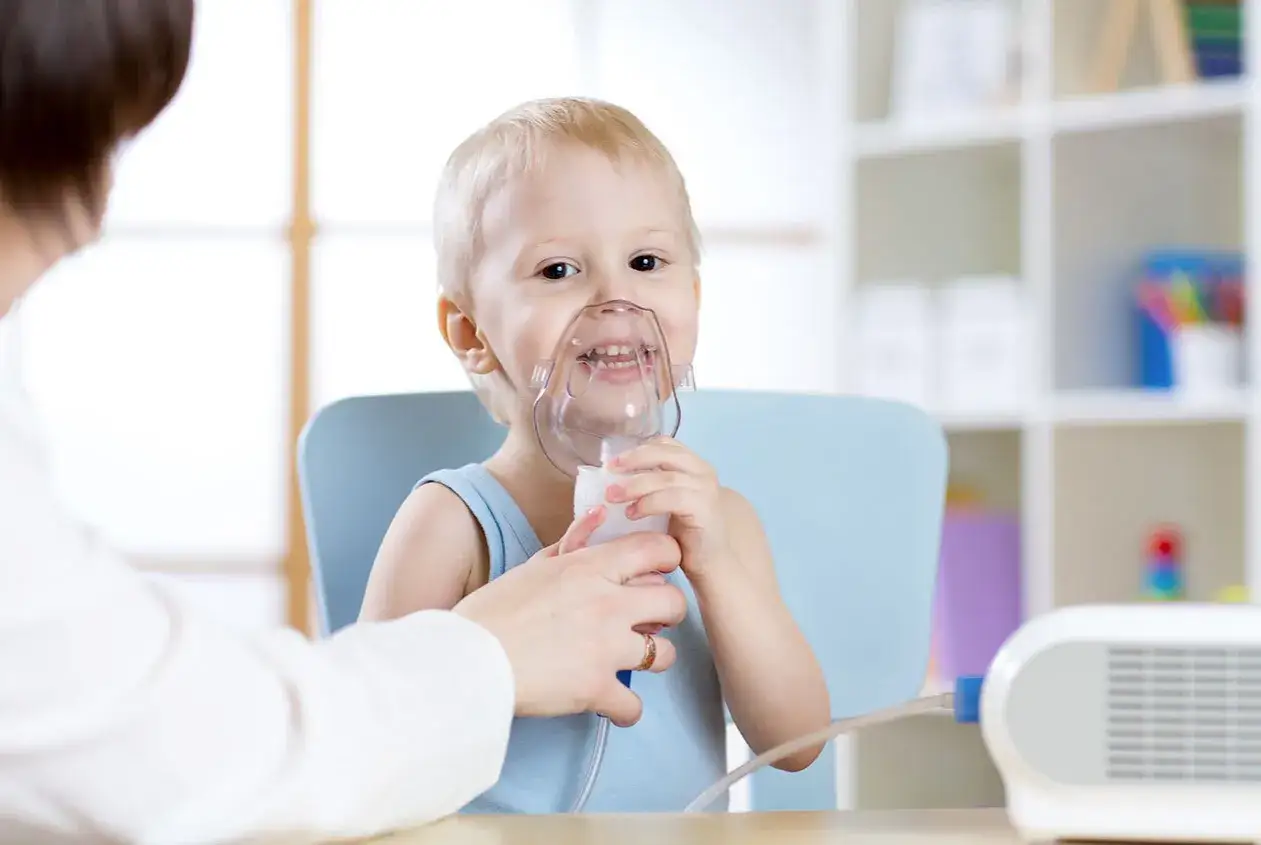 Bambino sorridente con maschera per aerosol, pronto per la terapia. La mamma lo assiste, assicurandosi che il trattamento sia fatto prima o dopo i pasti per il suo benessere.