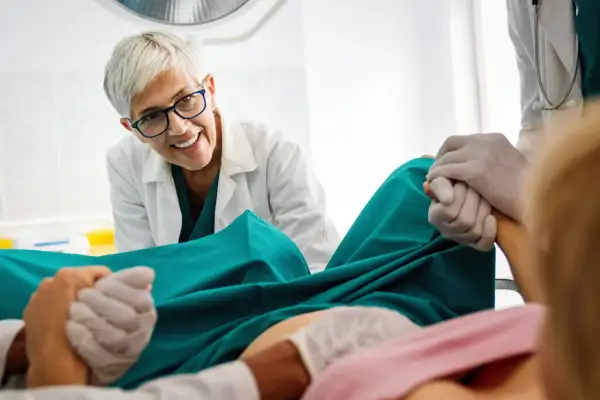 Medico sorridente assiste a un parto, tenendo la mano della paziente. Si percepisce un momento di scollamento membrane e attesa.