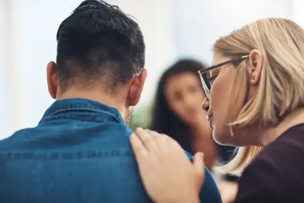 Una donna con gli occhiali consola un uomo, offrendo supporto per la psicosi da stress. Un'altra persona è sfocata sullo sfondo.