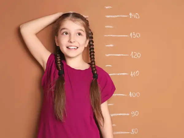 A young girl with braided hair smiles as she measures her height against a chalk growth chart.