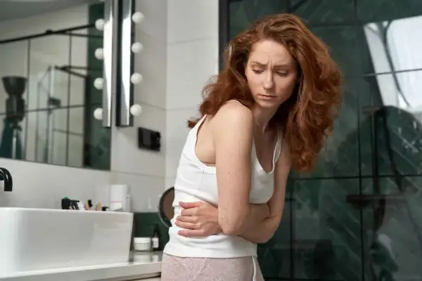 Donna con capelli rossi in bagno, stringe l'addome con le mani, visibilmente sofferente. Potrebbe essere un caso di diarrea prima del ciclo.